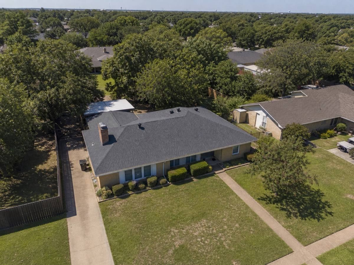Oblique aerial view of a mid-century brick ranch with two large roof planes and a rear patio cover, roofed in medium-charcoal architectural shingles, with neighbouring roofs and mature trees around it.