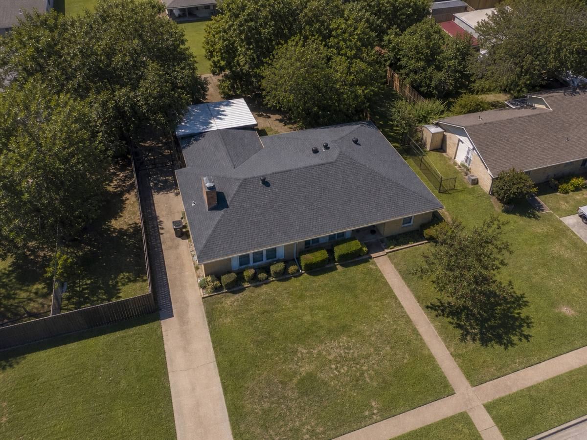 Overhead view looking straight down on a mid-century brick ranch with two large roof planes and a rear patio cover, showing the full run of medium-charcoal architectural shingles and how the planes drain.