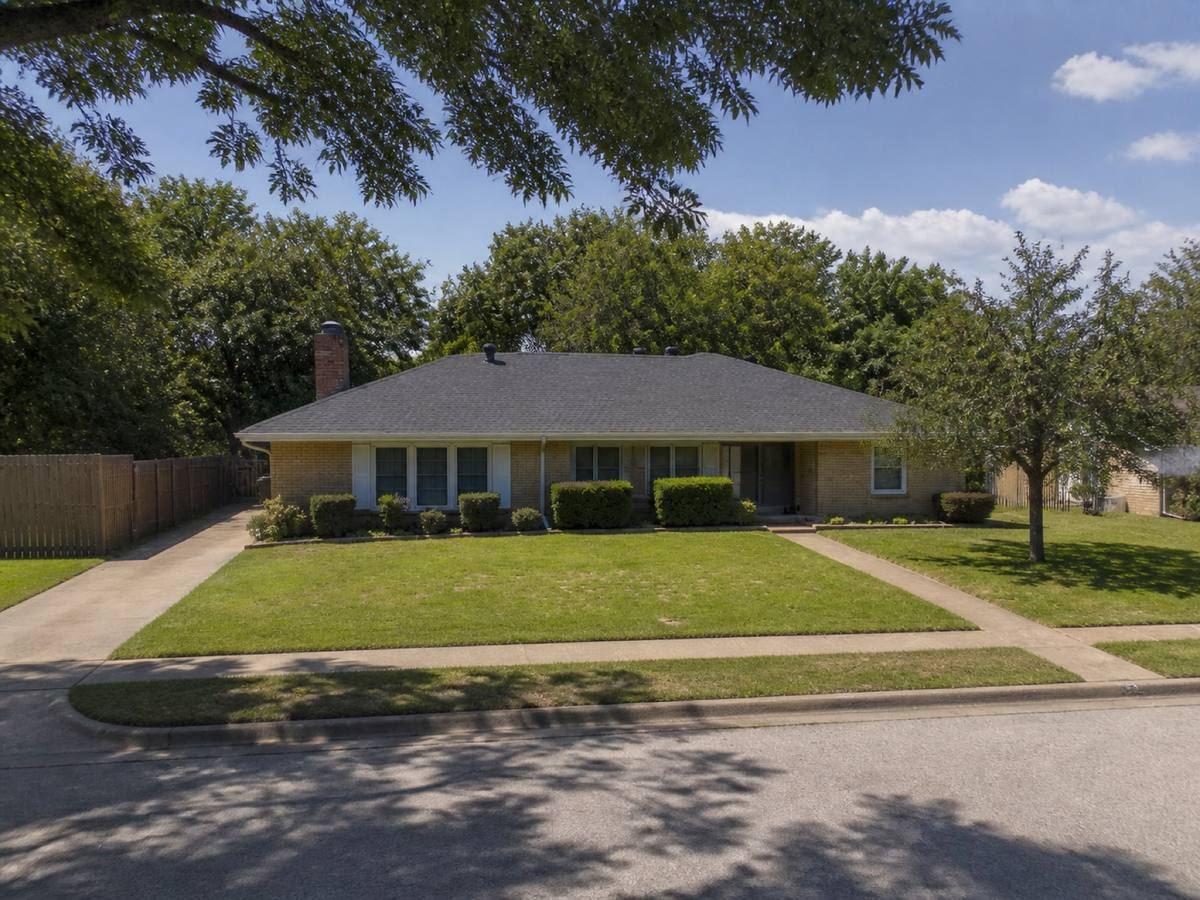 A single-storey tan-brick ranch house seen from the street beneath mature trees, its roof, fascia and front elevation in view.