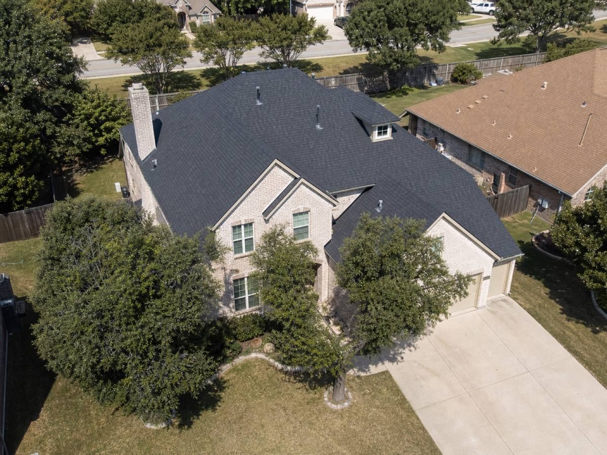 Oblique aerial view of a two-storey light-brick house with asymmetrical gables and a small rear dormer, roofed in dark slate dimensional shingles, with neighbouring roofs and mature trees around it.
