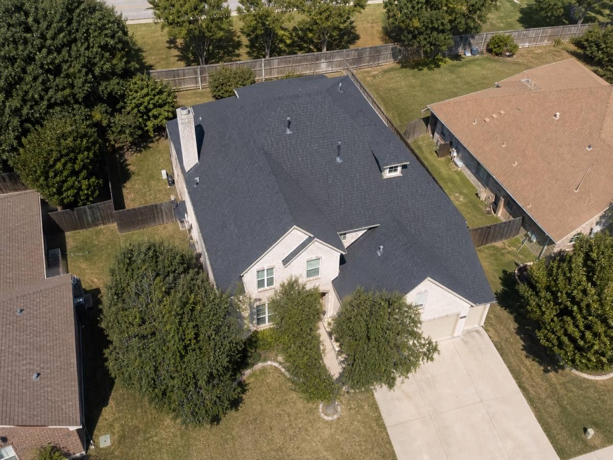 Overhead view looking straight down on a two-storey light-brick house with asymmetrical gables and a small rear dormer, showing the full run of dark slate dimensional shingles and how the planes drain.