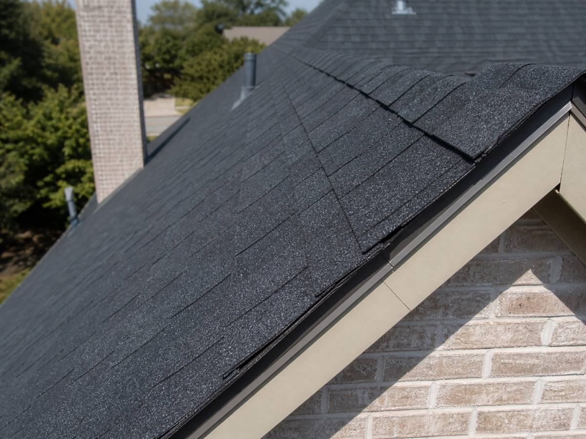 A dark shingle hip running down to a gable end, the drip edge and painted fascia visible along the rake above pale brick.