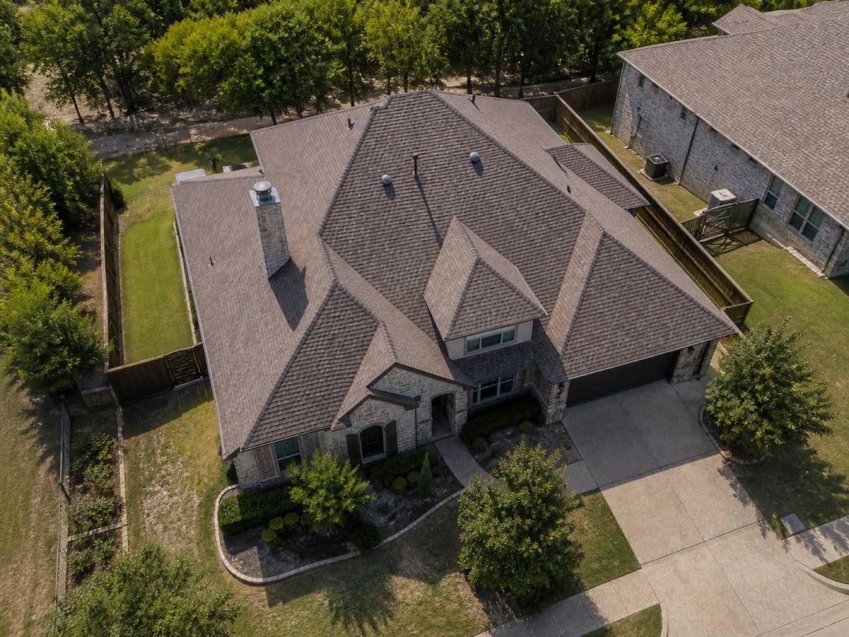 Overhead view looking straight down on a stone-and-brick house with a broad hip roof backing onto a greenbelt, showing the full run of weathered-brown dimensional shingles and how the planes drain.