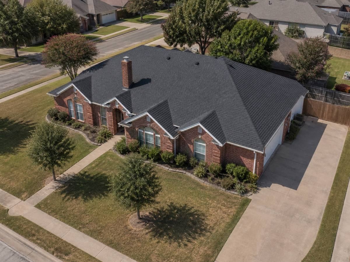 Oblique aerial view of a single-storey red-brick house with three front gables on a corner lot, roofed in charcoal impact-resistant shingles, with neighbouring roofs and mature trees around it.