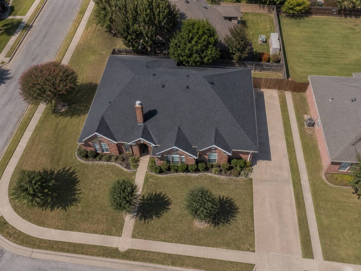 Overhead view looking straight down on a single-storey red-brick house with three front gables on a corner lot, showing the full run of charcoal impact-resistant shingles and how the planes drain.