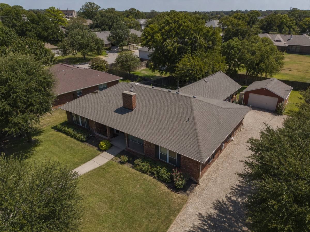 Oblique aerial view of a brick house with a long ridge and a detached workshop behind it, roofed in warm-grey architectural shingles, with neighbouring roofs and mature trees around it.