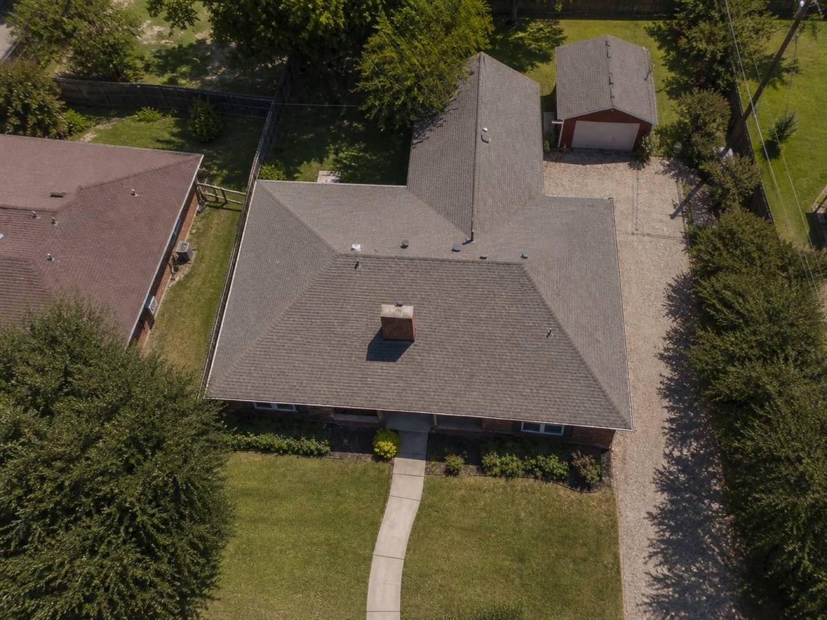 Overhead view looking straight down on a brick house with a long ridge and a detached workshop behind it, showing the full run of warm-grey architectural shingles and how the planes drain.