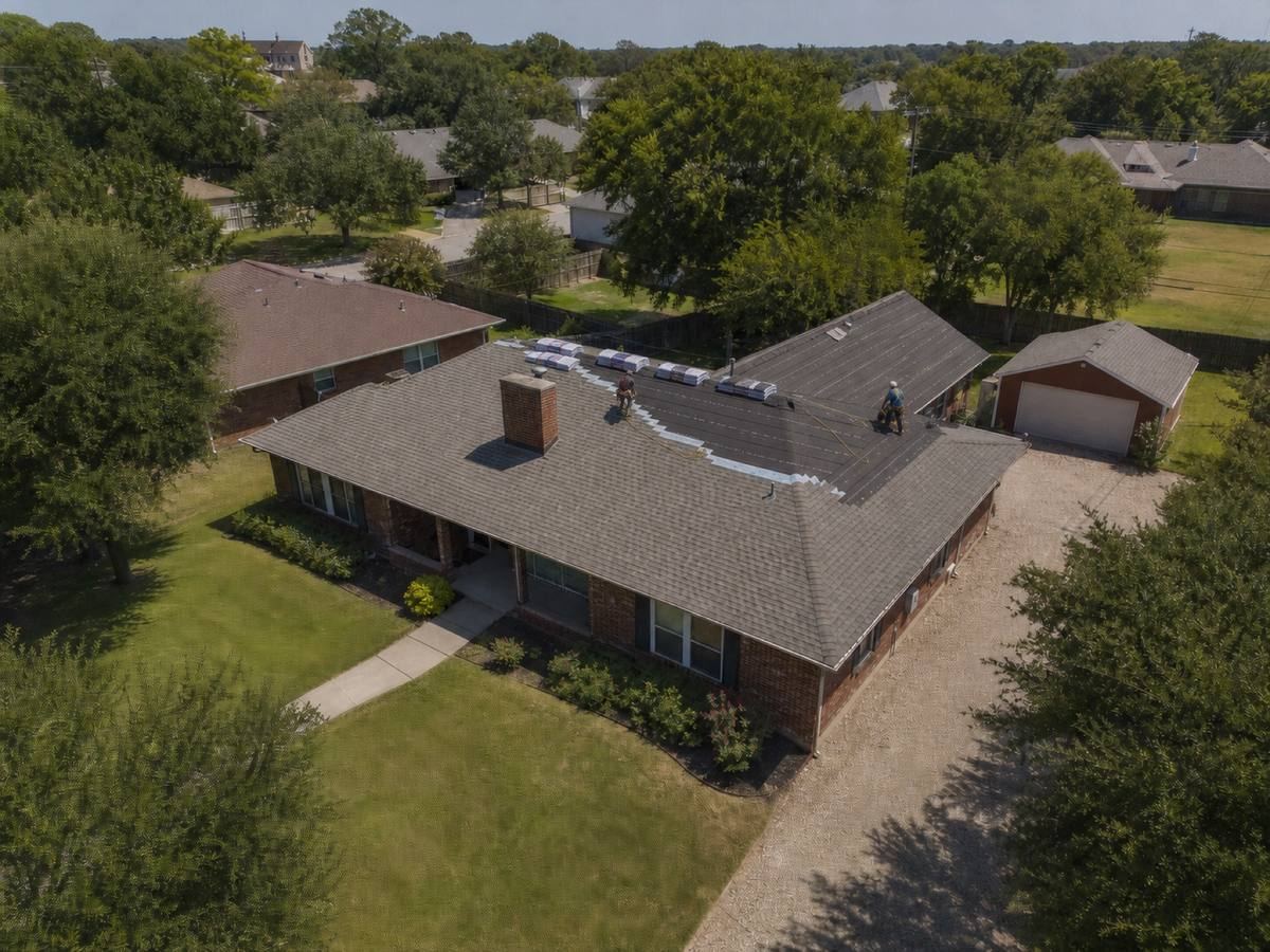 A brick house with a long ridge and a detached workshop behind it part-way through a architectural shingle installation, with finished and unfinished areas both visible.