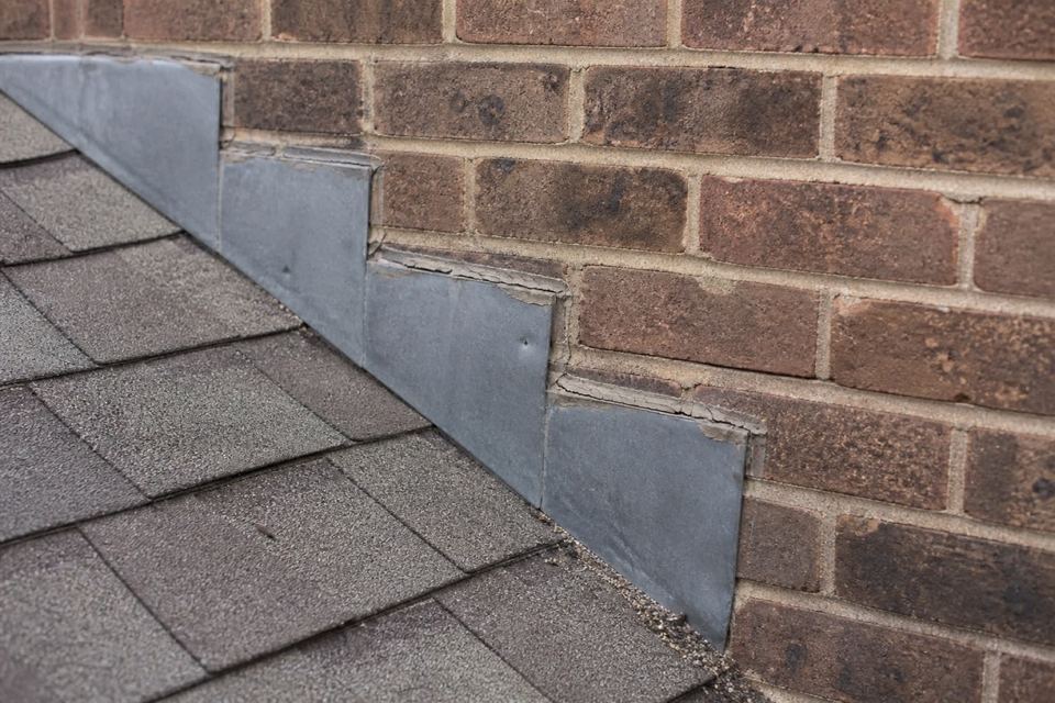 Step flashing where a shingle plane meets a brick wall, an old sealant bead cracked along the top edge.